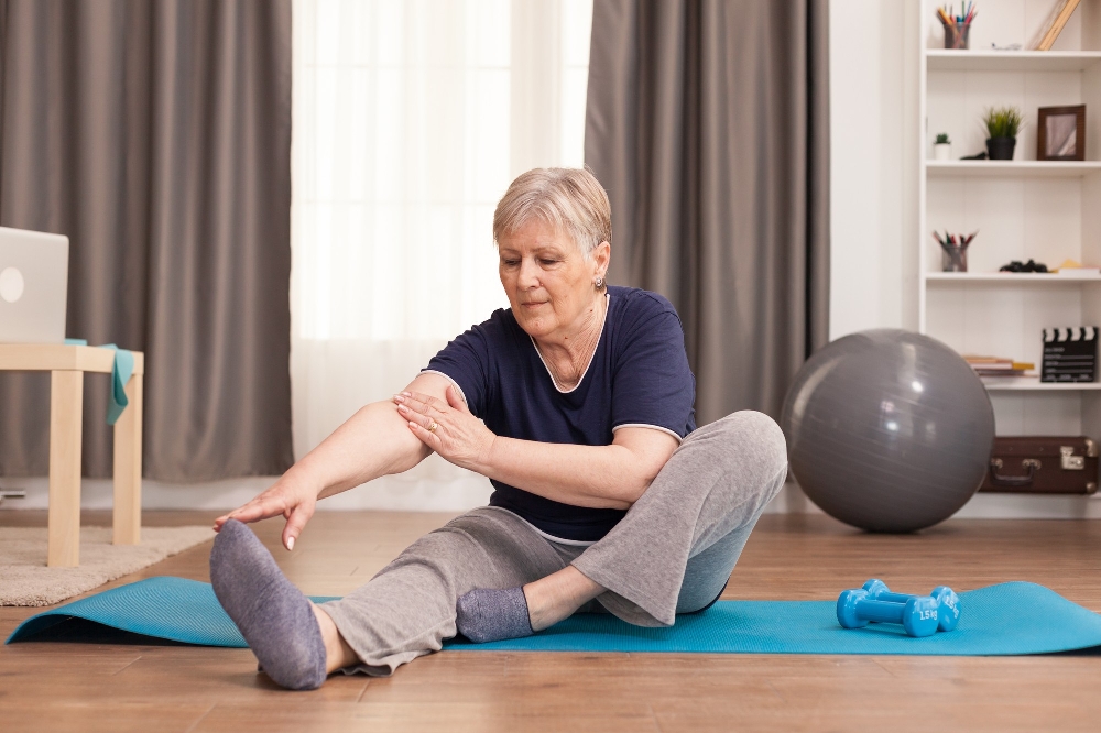 Older woman stretching toward her foot on a teal mat at home, a gentle movement scene for living with rheumatoid arthritis